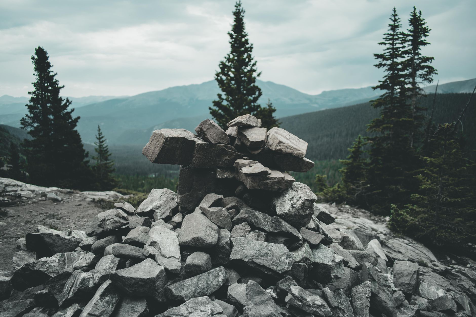 A stack of rocks amidst a dramatic mountain landscape with evergreen trees and cloudy skies.